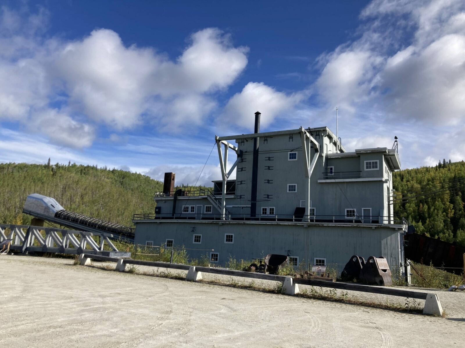 A gold dredge with trees behind it. The dredge is four stories high and made of wood painted gray. There. is a large bucket line coming out of one end.