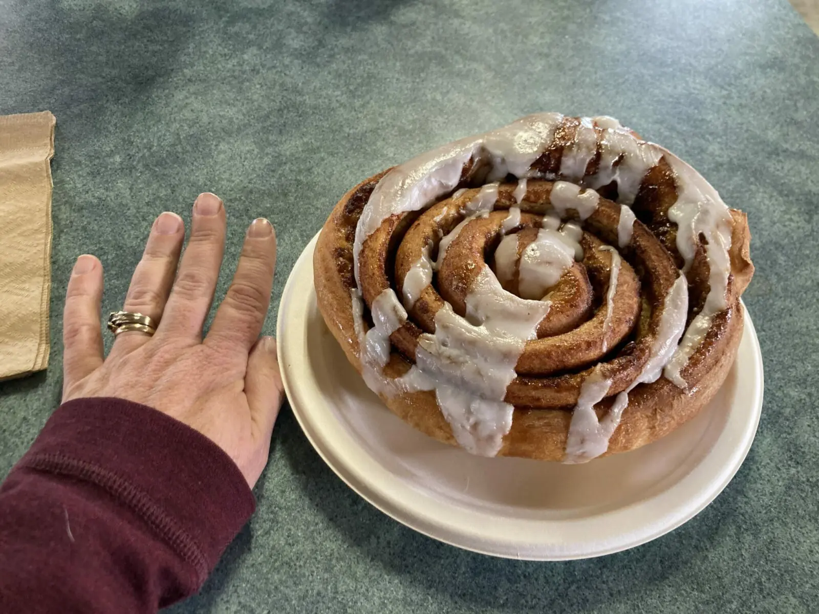 A large cinnamon roll covered in frosting with a person's hand for scale