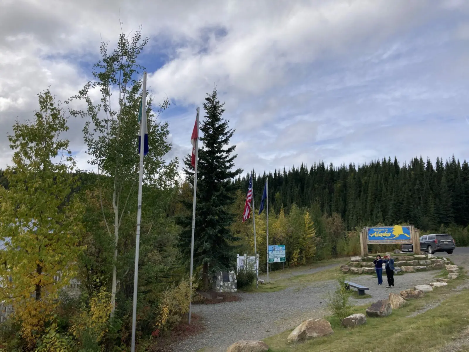 The border between the US and Canada and Alaska and Yukon. There is a welcome to Alaska sign and several flags and people taking photos.