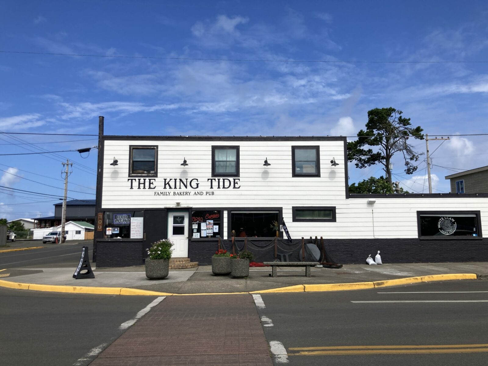 A white painted wooden building with black trim on  a street corner. The sign says King Tide Family bakery and pub