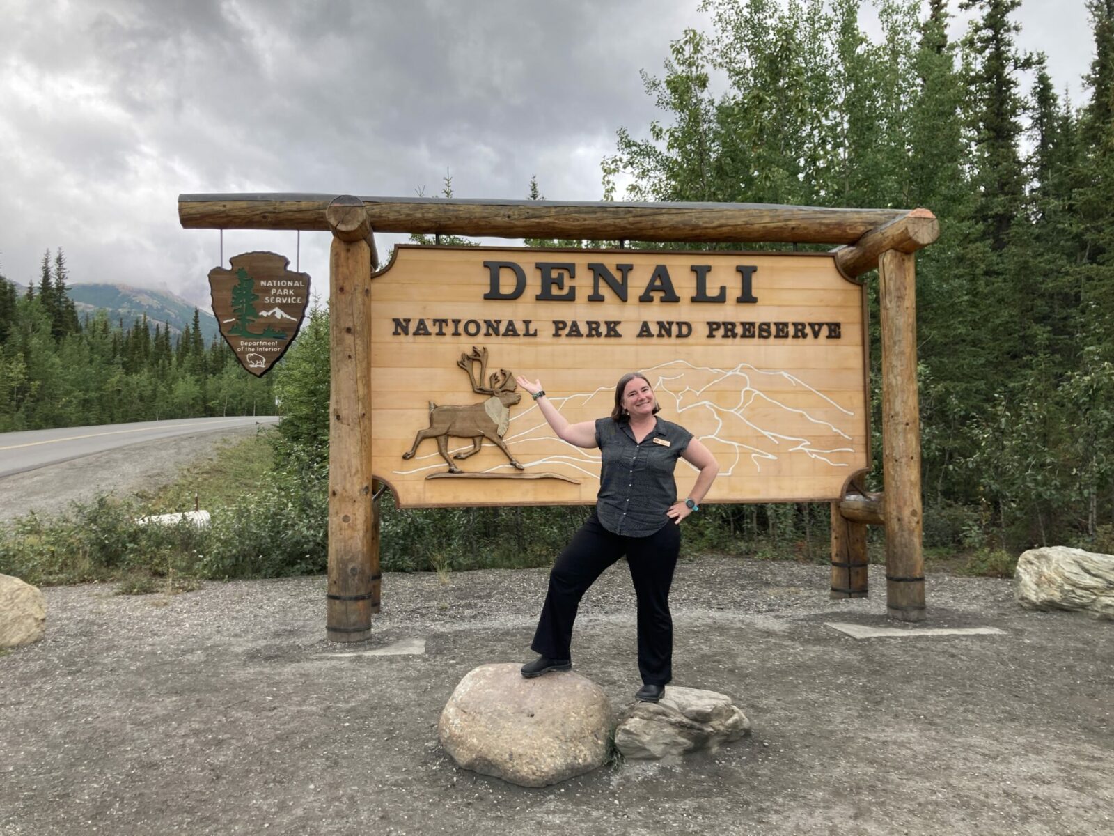 Jennie in front of the Denali National Park and Preserve sign, a large wooden sign in the forest next to the road with a caribou and mountains and the National park service logo