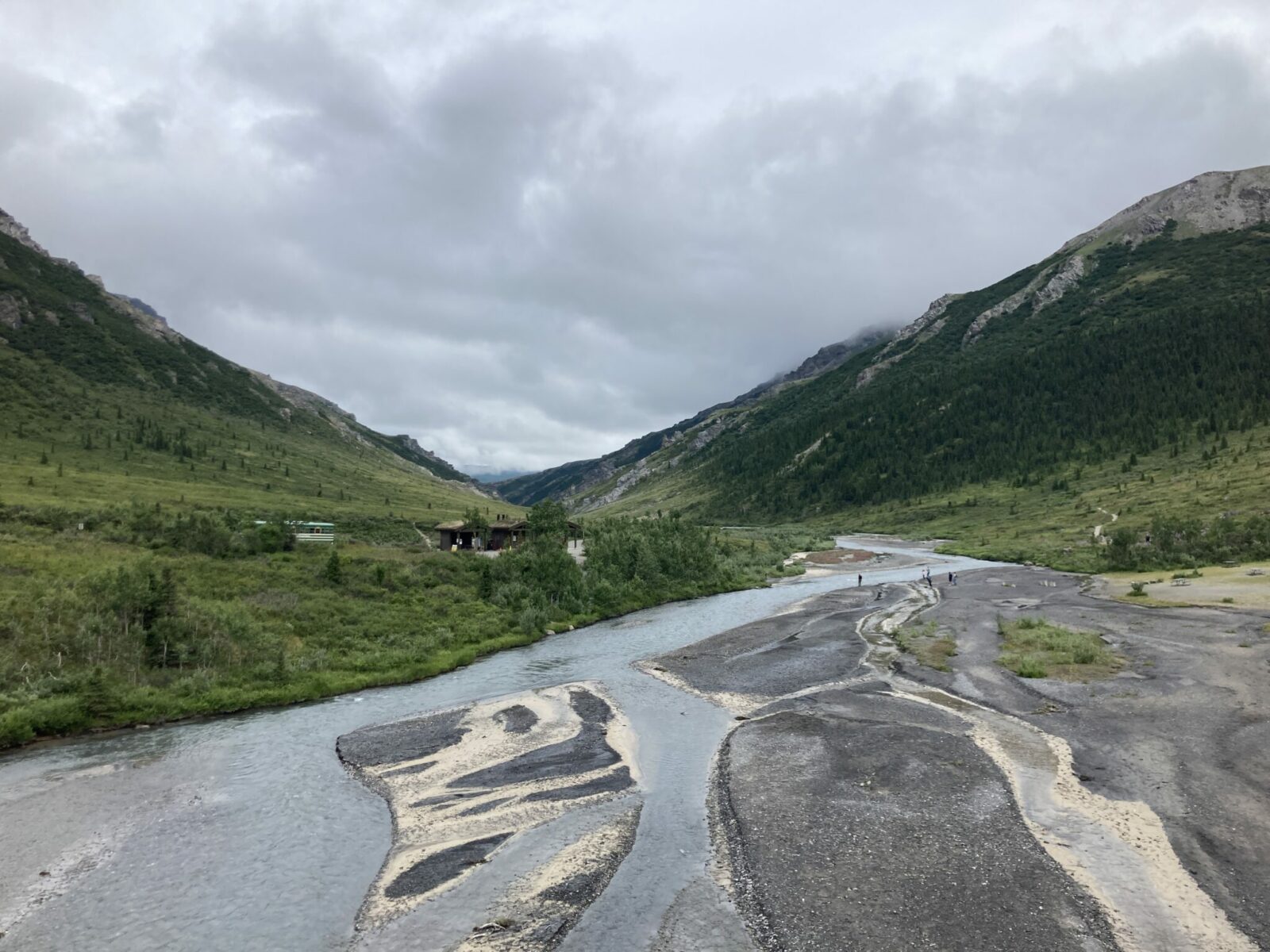 A bradied gravel river between forested mountains on a cloudy day. A few hikers are on the gravel near the river