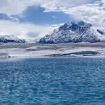 A blue pool of water on Knik Glacier surrounded by white and gray ice and high snow covered and glaciated mountains