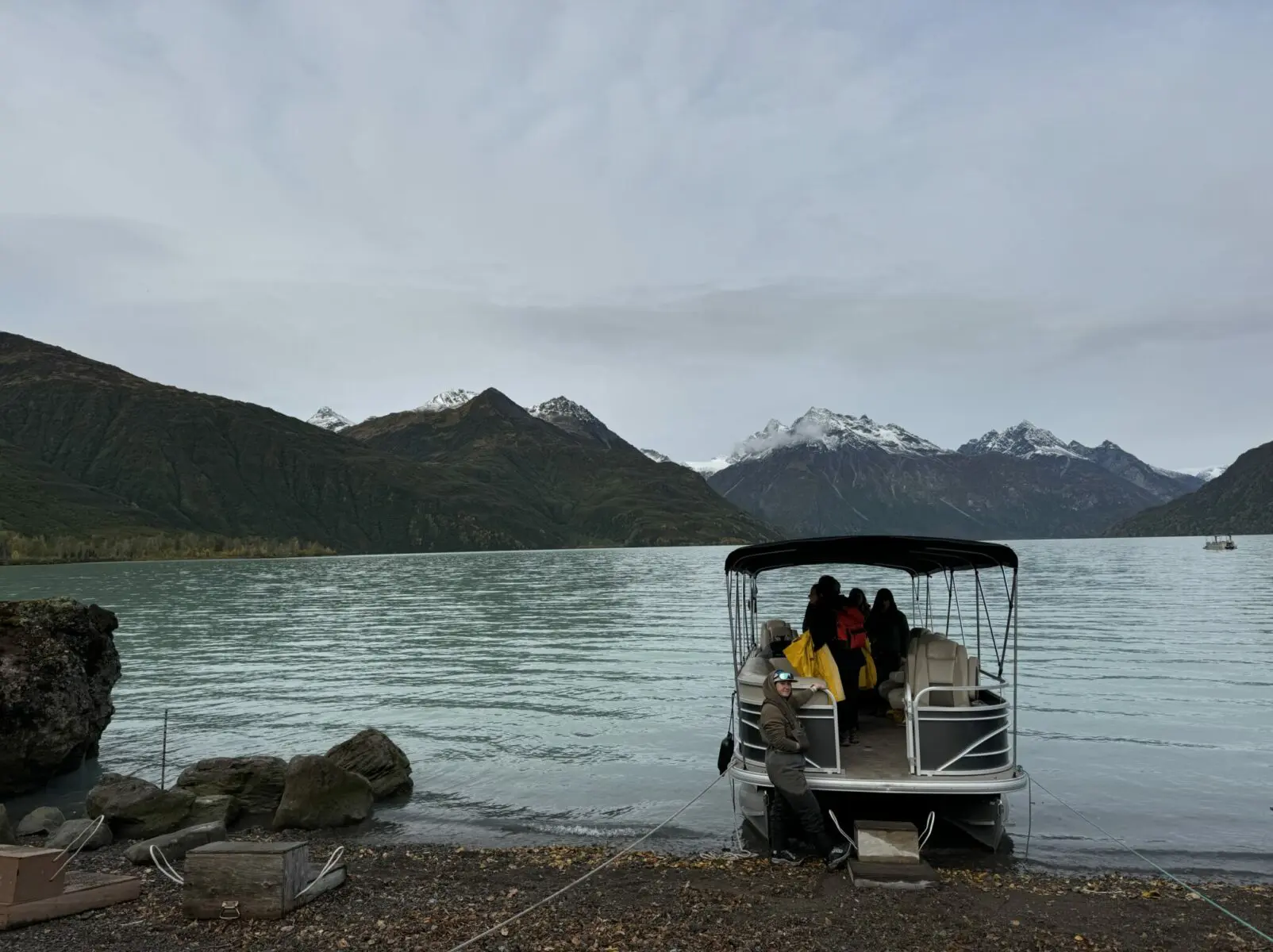 A pontoon boat at the edge of a lake tied to the shore with a wooden step for guests to get on board. Mountains with fresh snow are in the background on an overcast day