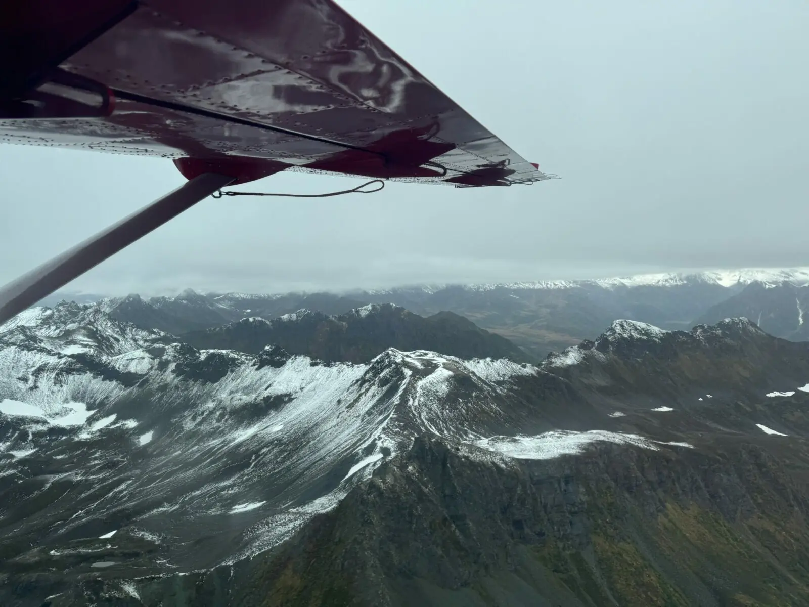A red airplane wing of a small plane on an overcast day with mountains with fresh snow below