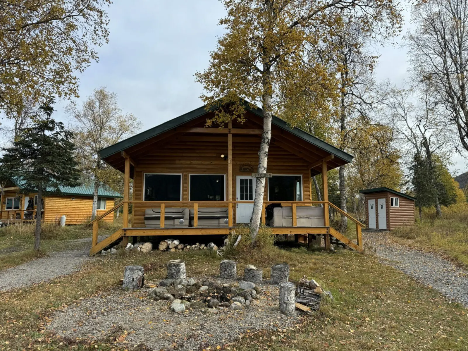 A log building with seating on the porch and a firepit in the foreground. There is an outhouse and another cabin behind the building and several golden birch trees are around the buildings.