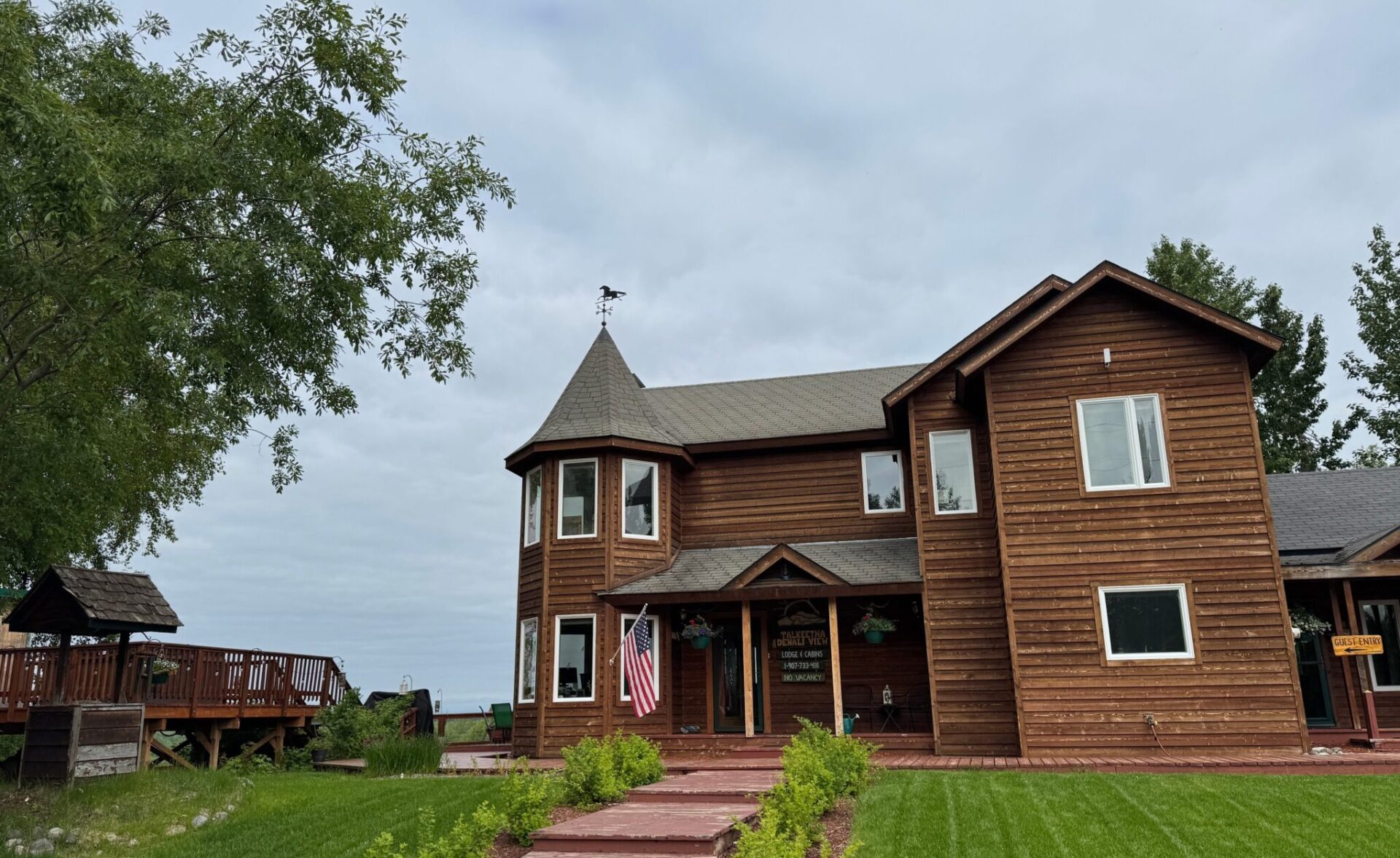 A photo of Talkeetna Denali View Lodge showing a walkway to the front, a wood building with a turret and a deck around the back